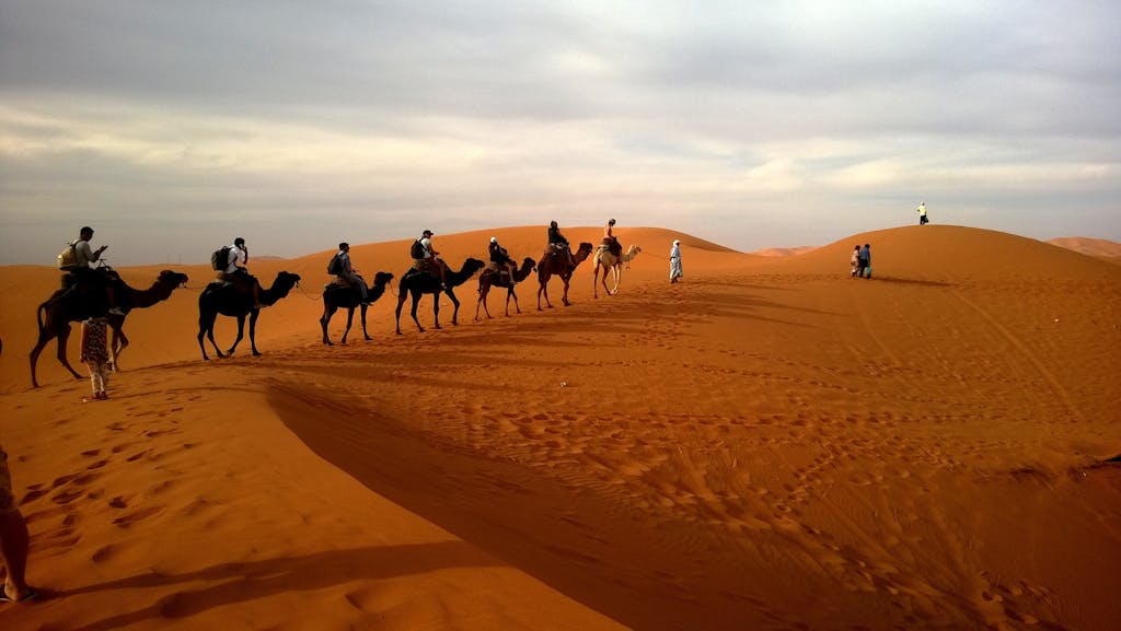 A group traveling on camels over reddish sands under a cloudy sky in a vast desert landscape.