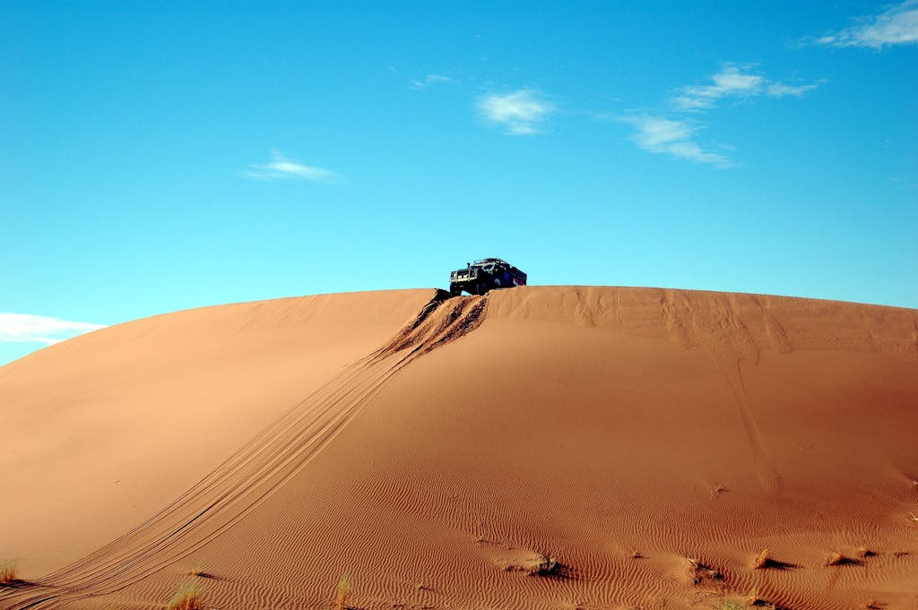 A rugged off-road vehicle climbs a vast sandy dune under a clear blue sky.